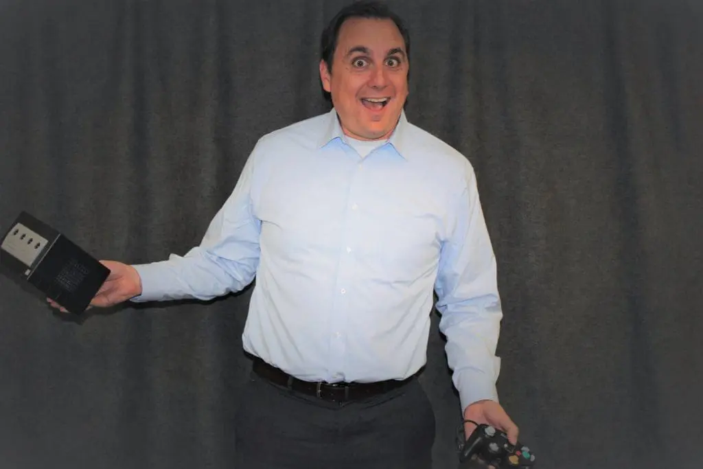Jake Slayton in a light blue shirt smiling broadly and holding a video game console and controller against a gray background.