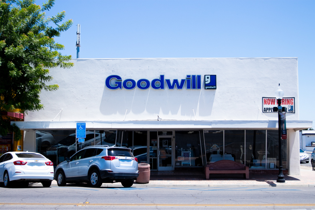 Goodwill Delano thrift store exterior with cars parked in front, a tree on the left, and a "Now Hiring Apply Inside" sign on a lamp post.