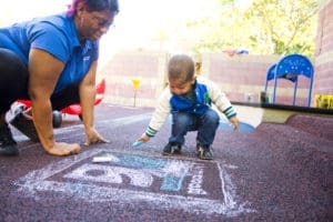 A woman and a toddler draw with chalk on the ground at a playground. The toddler is squatting and holding a piece of chalk while the woman is kneeling beside him, both focused on the drawing.