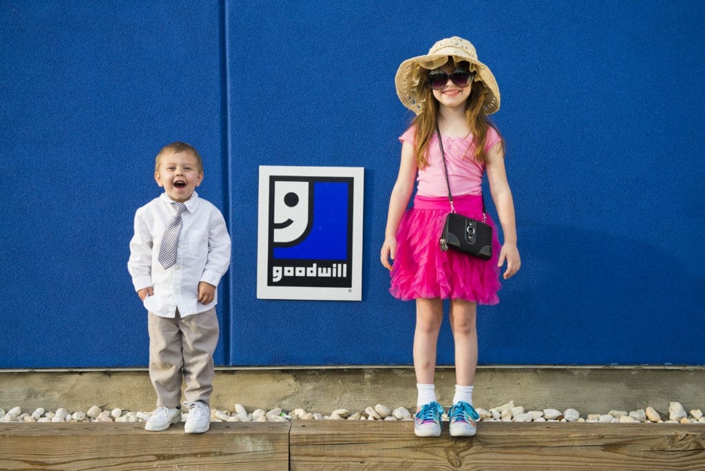 Two children are standing in front of a blue wall with a Goodwill sign. One child wears a shirt, tie, and pants, smiling widely. The other child wears sunglasses, a sunhat, pink shirt, and skirt, smiling.