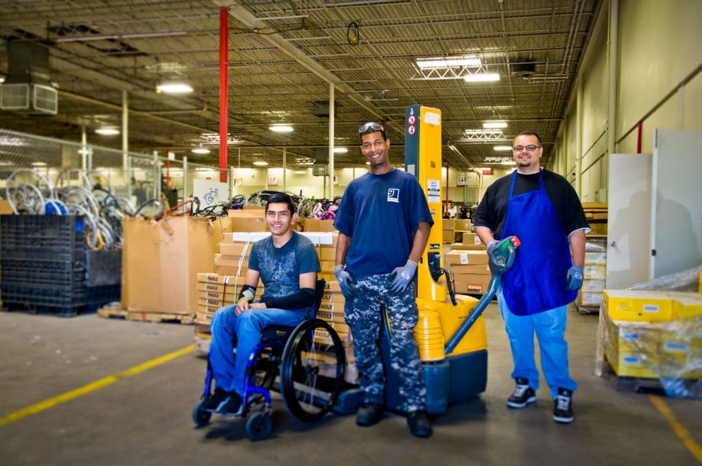 Three people posing in a warehouse with various boxes and materials in the background. One person is in a wheelchair, while the other two stand beside a yellow pallet truck.