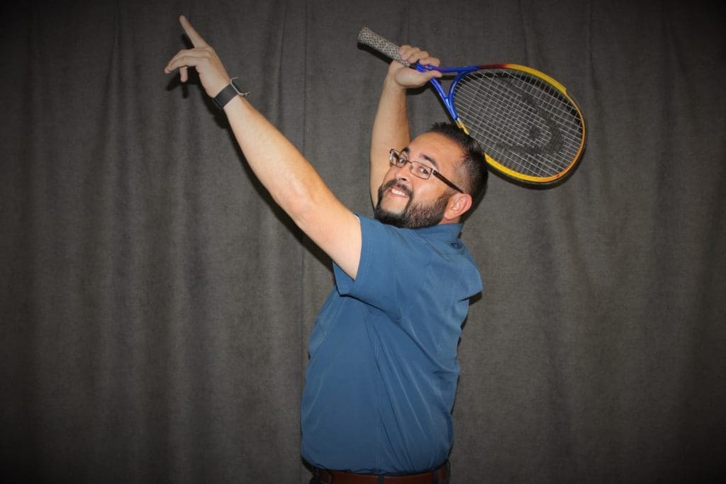 Justin Ahart wearing glasses and a blue shirt holds a tennis racket above his head while pointing upwards with his other hand, standing against a dark gray background.