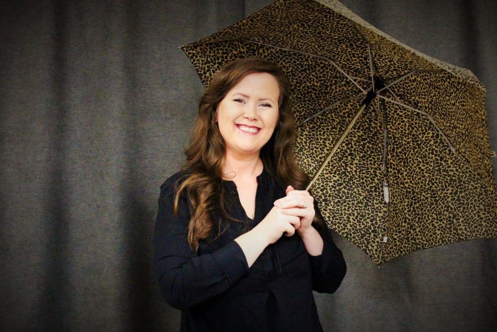 Randi Quicksall with long brown hair and a black outfit smiles while holding a leopard-print umbrella against a gray background.