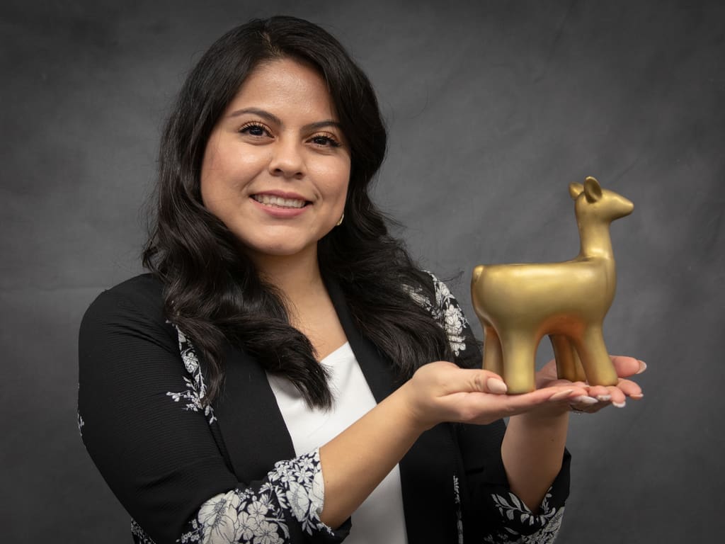 Brianda Ibarra with long dark hair, wearing a black and white outfit, smiles while holding a small golden figurine of a llama.