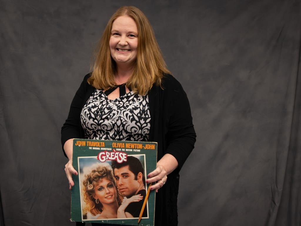 Rachel Moreno with shoulder-length hair holds a "Grease" vinyl record cover featuring Olivia Newton-John and John Travolta against a plain gray backdrop.