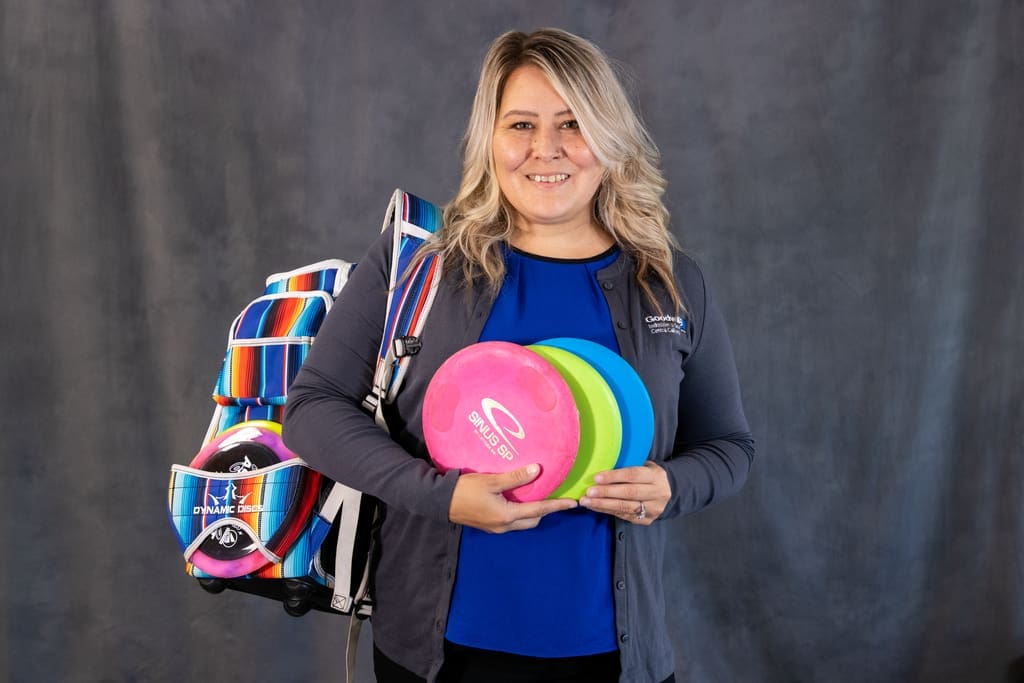 Jillian Mercer with long hair holding four disc golf discs is standing in front of a dark backdrop, with a colorful striped bag over their shoulder.