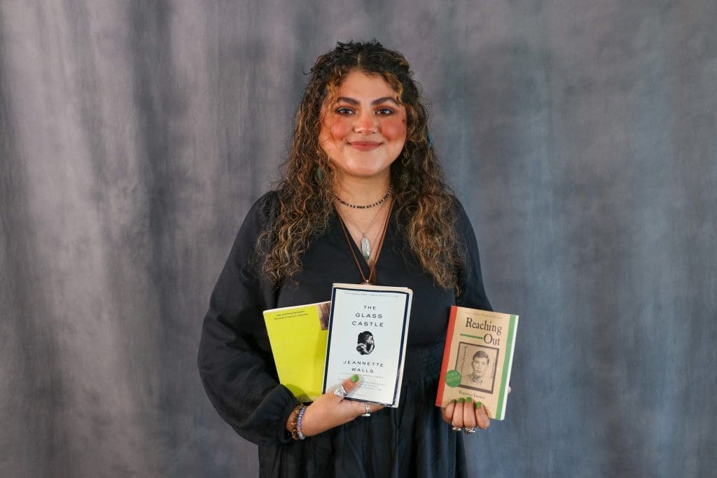 Marian Nasvid Torres with curly hair and a black dress holds three books: a yellow one, "The Glass Castle" by Jeannette Walls, and "Reaching Out" by Francisco Jiménez. They stand against a gray background.