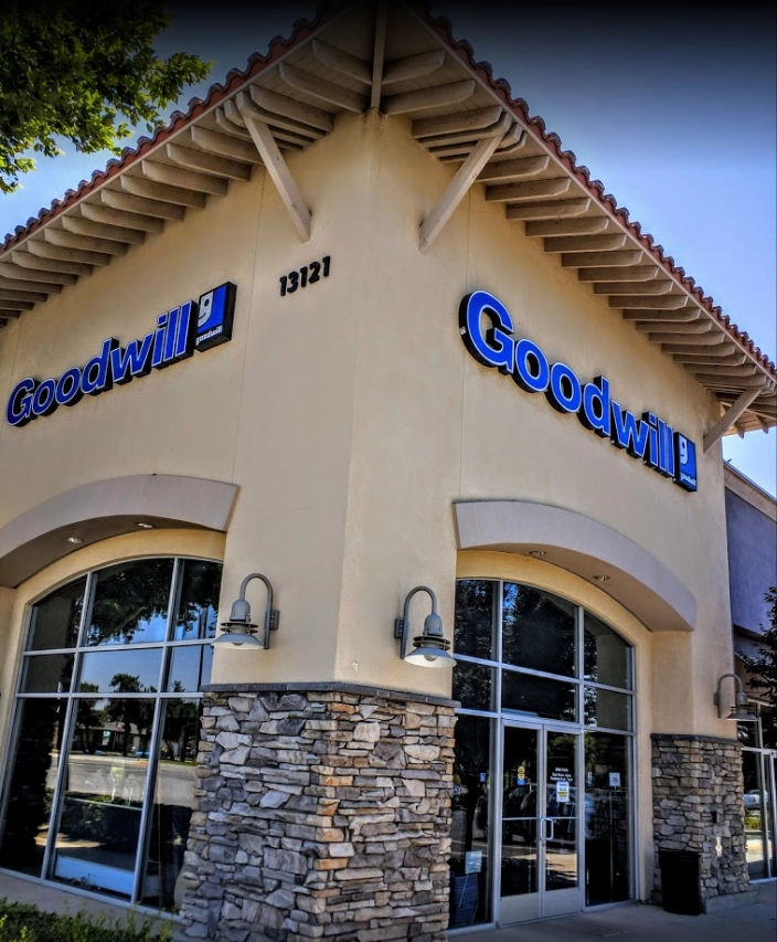 Front exterior of a Goodwill store with stone and stucco facade, large blue and white signage, and arched windows and doors. Address "13121" visible above entrance.