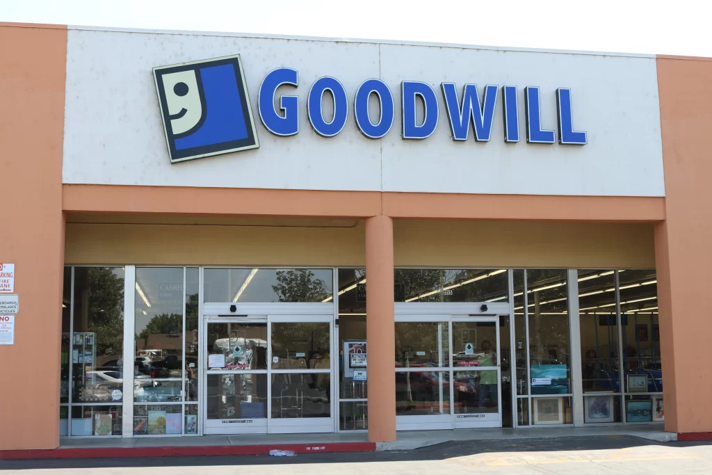 Exterior of a Goodwill store with large glass doors and windows displaying items inside. The store sign features the Goodwill logo and name above the entrance.