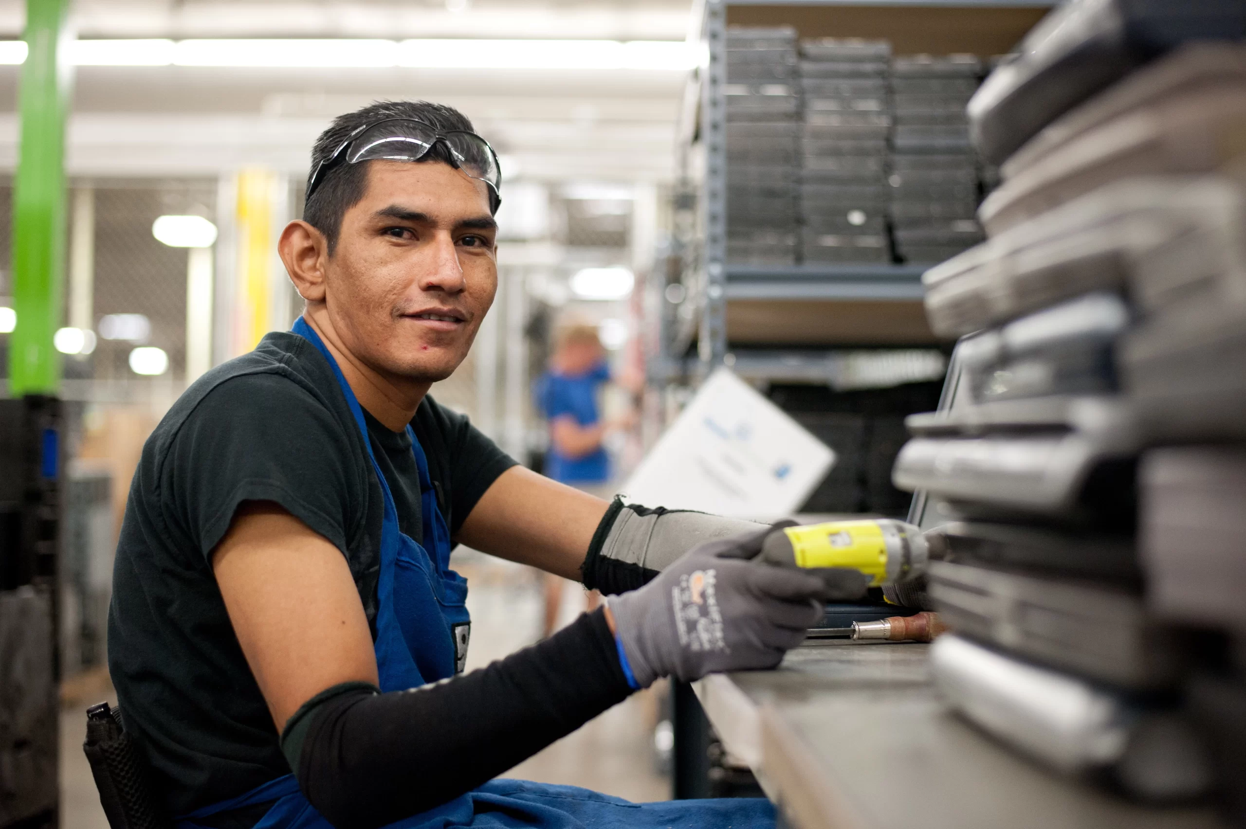 A worker wearing a blue apron and protective gloves uses a drill while assembling parts on a production line in a factory.