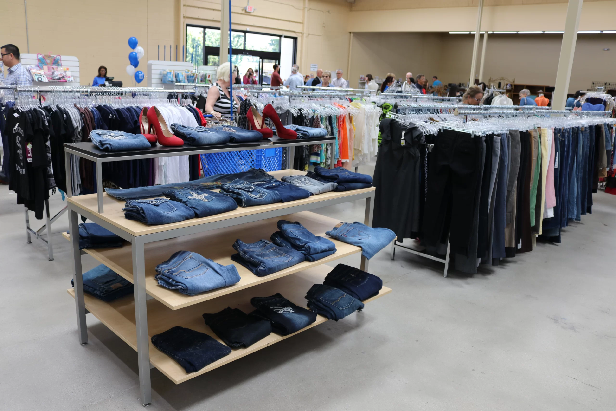 A clothing store with display tables showcasing folded jeans, shelves with red high-heel shoes, and racks of various clothing items. Shoppers browse in the background.