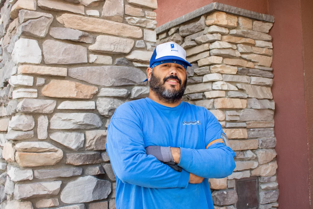 Man in a blue long-sleeve shirt and cap standing with arms crossed in front of a stone wall.