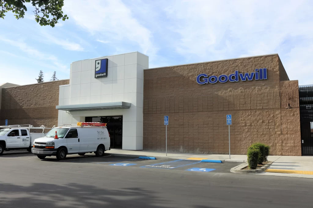 Exterior of a Goodwill store with a white van parked in front, accessible parking spaces, and a tree branch partially visible at the top left.