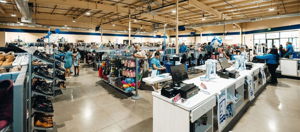 A spacious retail store interior with numerous customers browsing clothing racks and store associates assisting at busy checkout counters. The store has a high ceiling and bright lighting.