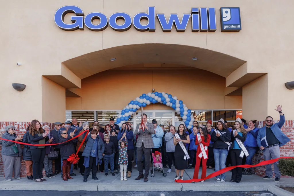 A group of people stands in front of a Goodwill store during a ribbon-cutting ceremony, with blue and white balloons decorating the entrance.